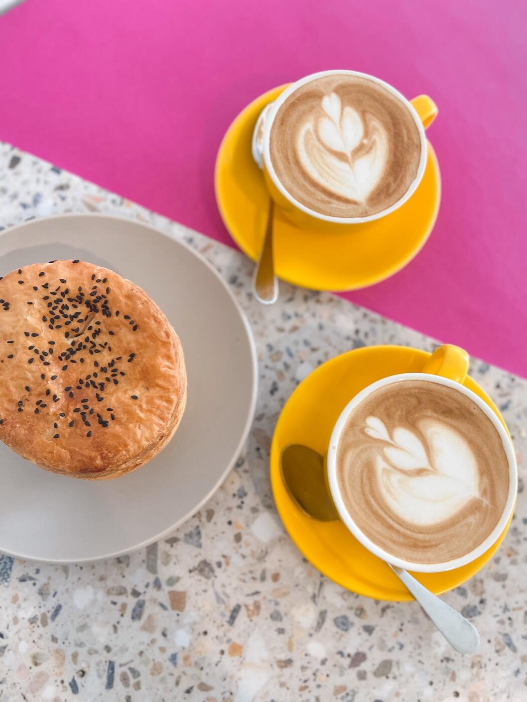 Two cappuccinos with latte art in yellow cups beside a round pie topped with black seeds, seen from above on a speckled benchtop
