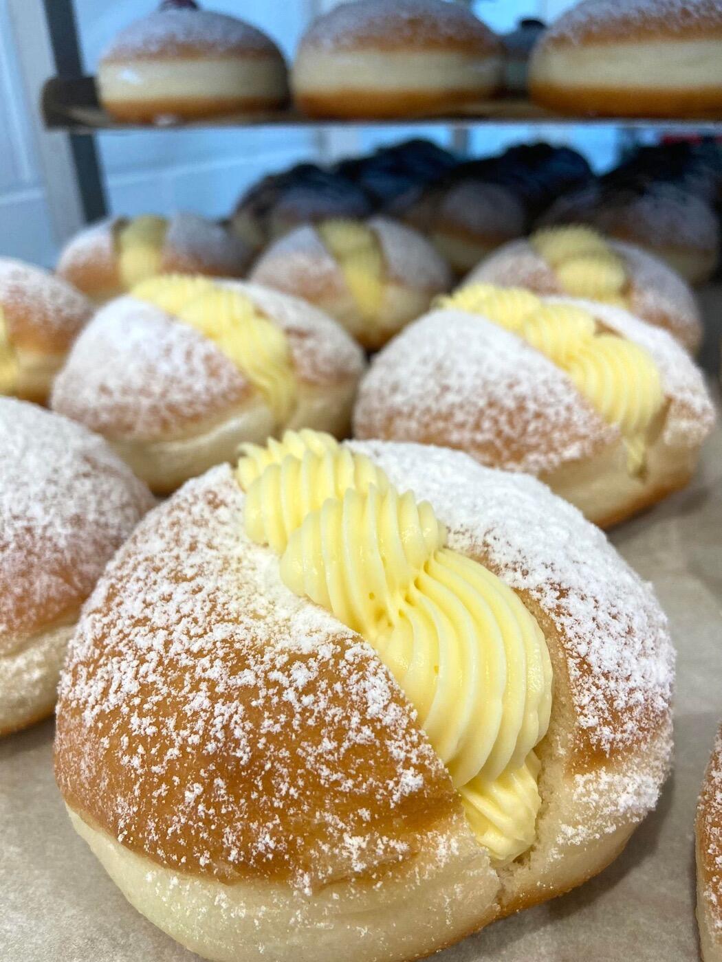 Round donuts dusted with icing sugar and topped with a swirl of yellow cream, with more donuts on the rack behind