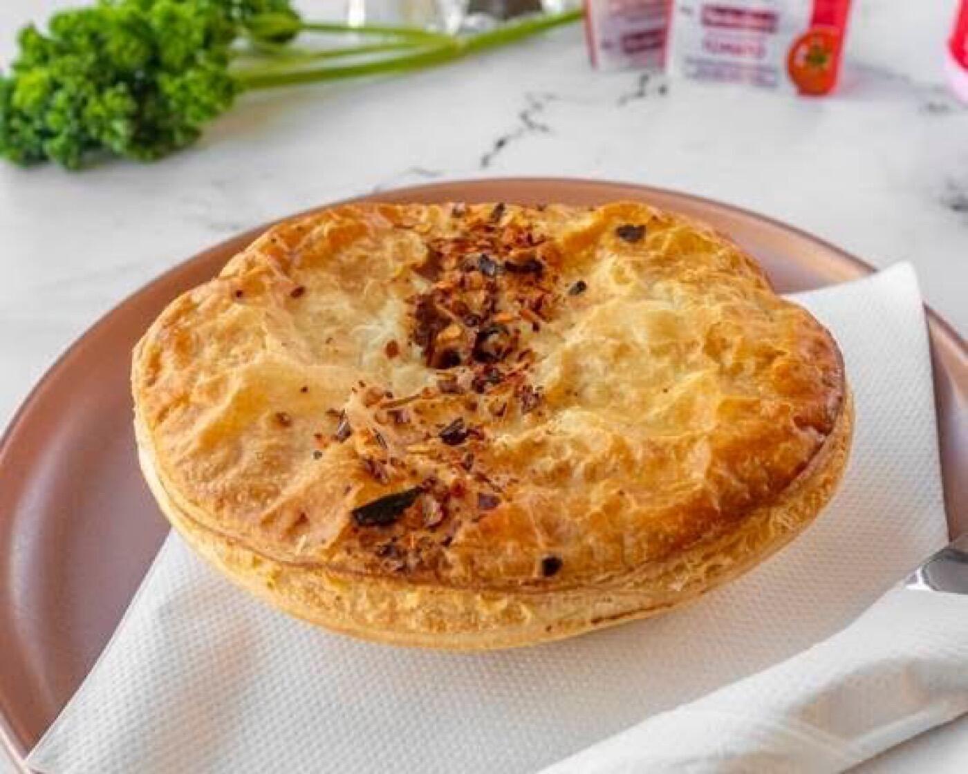 A golden pie with a crisp, flaky pastry lid flecked with dark crumbs along the top, on a pink plate with a paper napkin