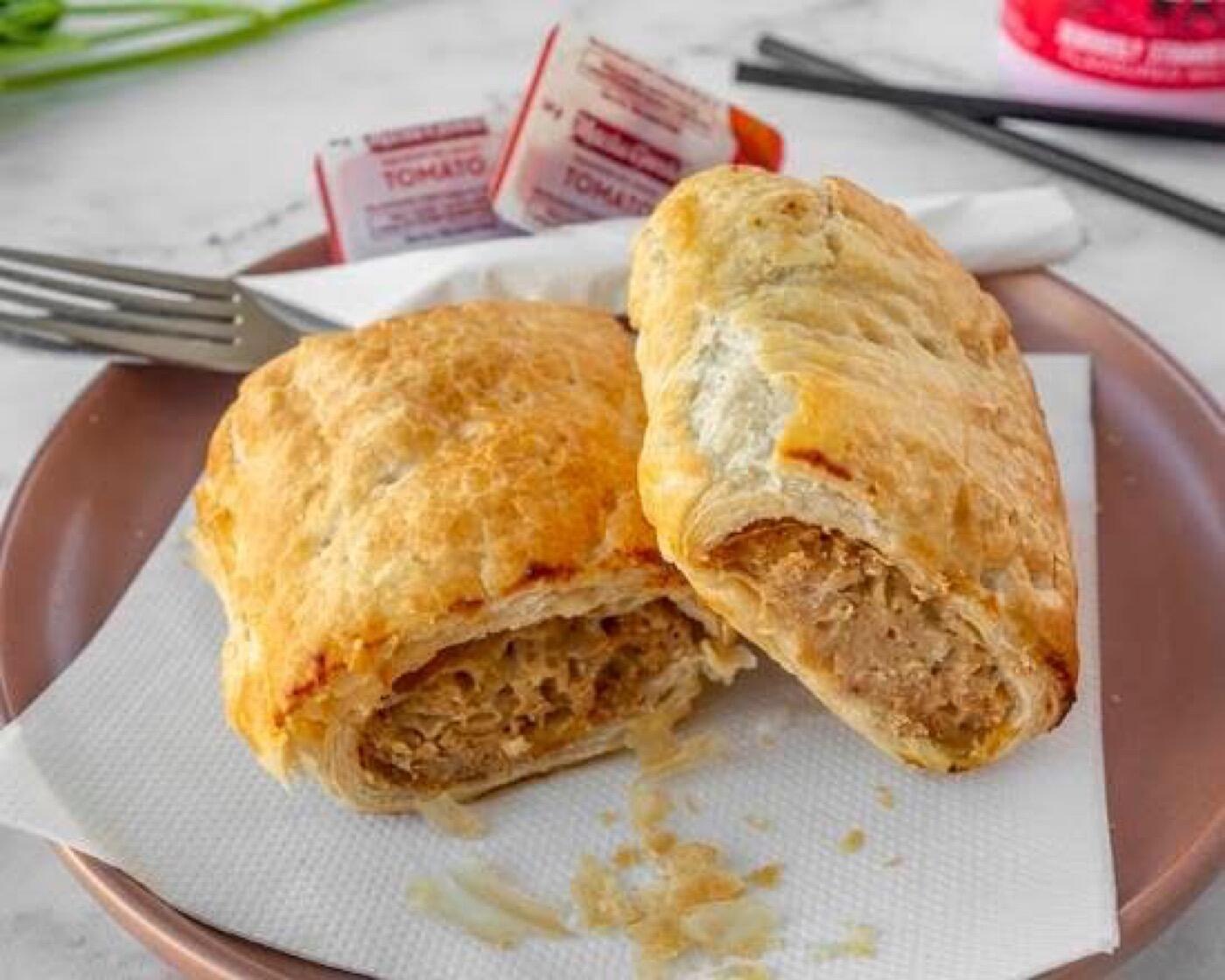 A sausage roll cut in half to show the filling inside golden flaky pastry, on a pink plate