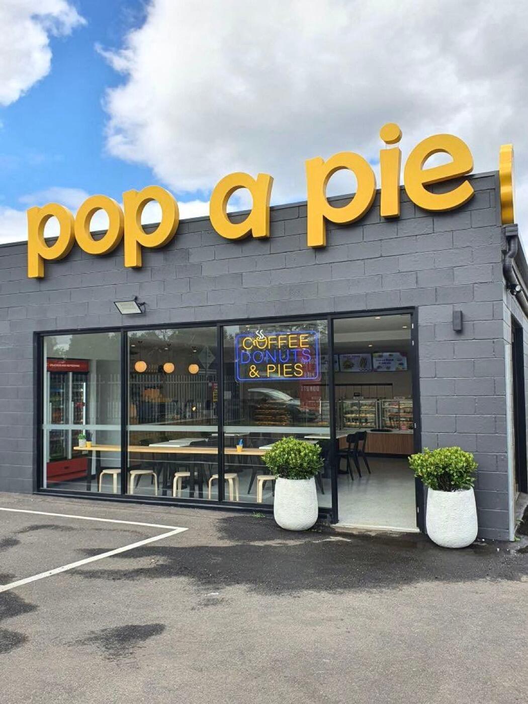 The grey brick shopfront with a yellow 'pop a pie' sign above big glass windows and two white planters by the door